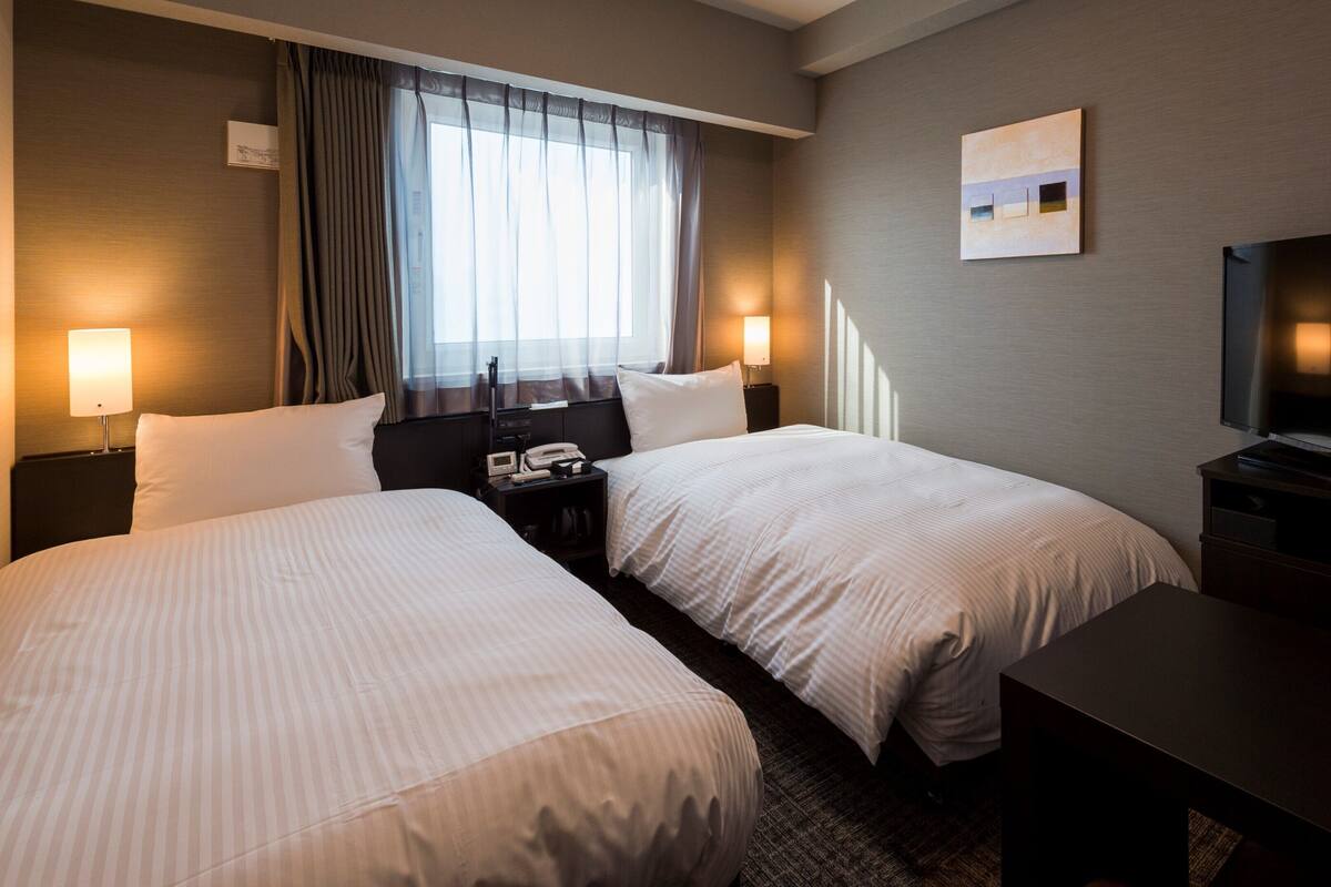 A hotel room with two beds, a TV, a desk, and a window with curtains.