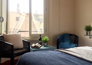 Un dormitori amb un llit, dues cadires, una taula petita amb una ampolla i una planta, i una finestra que dona vistes a edificis.
