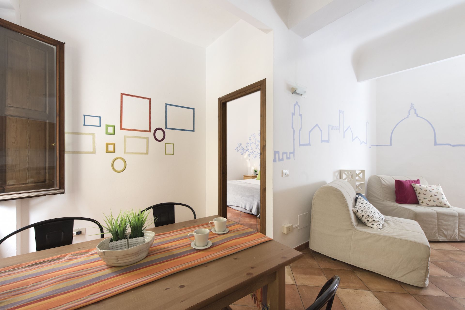 Una sala de estar acogedora con una mesa de comedor de madera, un sofá y una pared decorada con formas geométricas y un mural con vista a la ciudad.