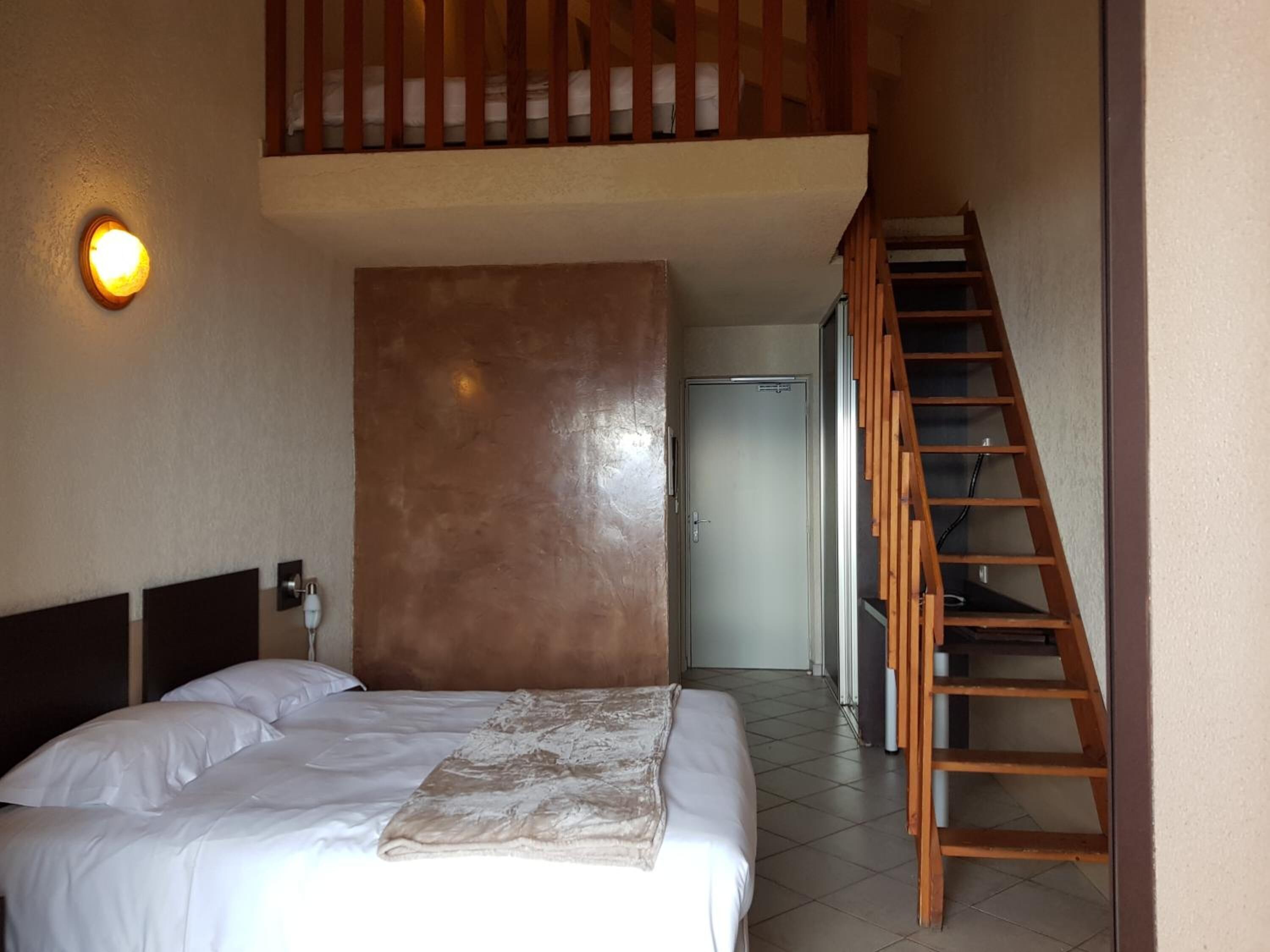 Un dormitorio con cama, cabecera de madera, mesita de noche, lámpara de pared, un espejo grande y una escalera de madera.