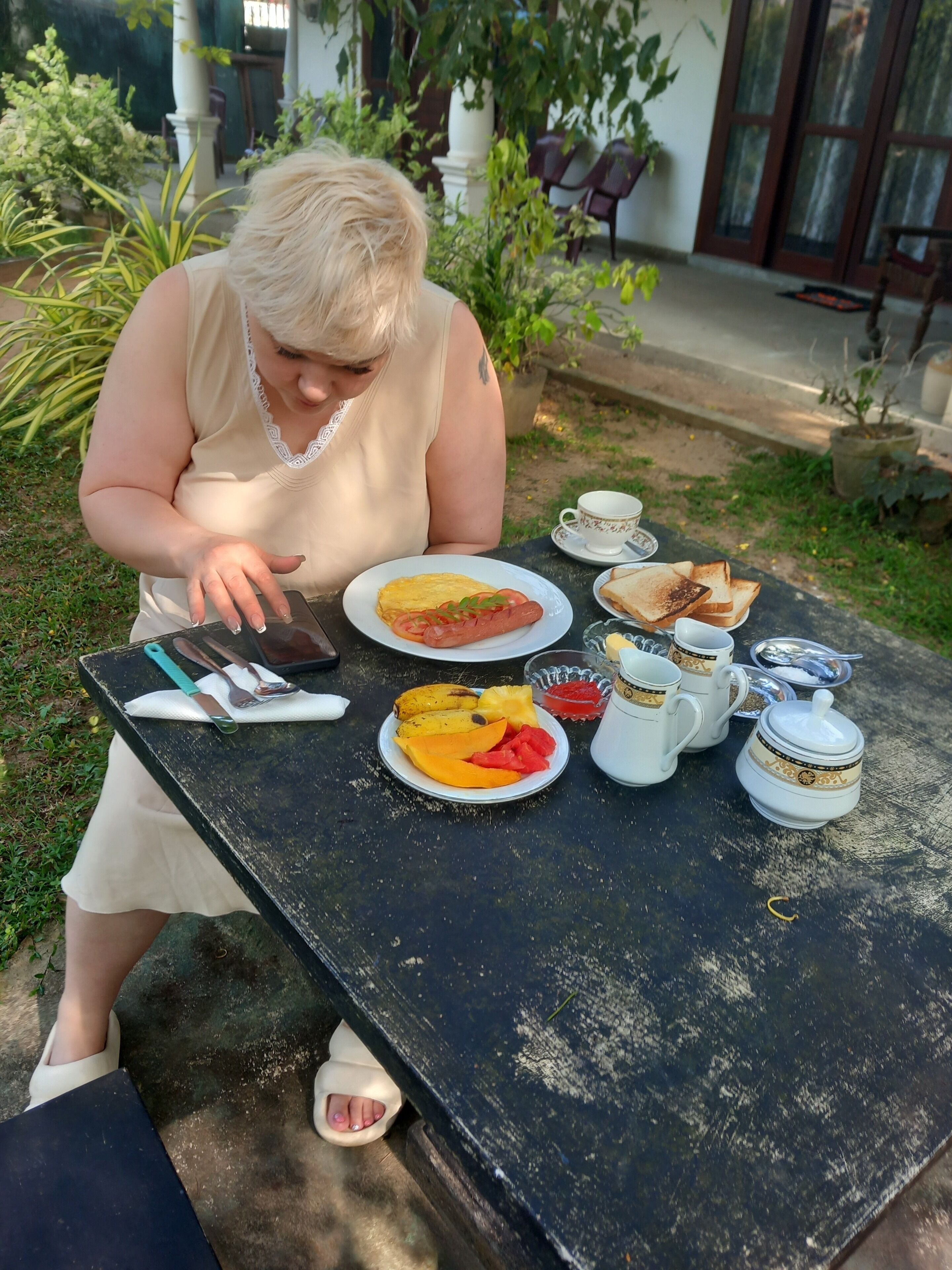 Una persona sentada en una mesa al aire libre con un plato que incluye tostadas, fruta y un hot dog. Hay tazas y una tetera sobre la mesa.