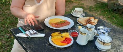 Una persona sentada en una mesa al aire libre con un plato que incluye tostadas, fruta y un hot dog. Hay tazas y una tetera sobre la mesa.