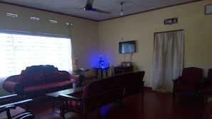 A living room with a red sofa, wooden chairs, a table, a TV on the wall, and a ceiling fan.