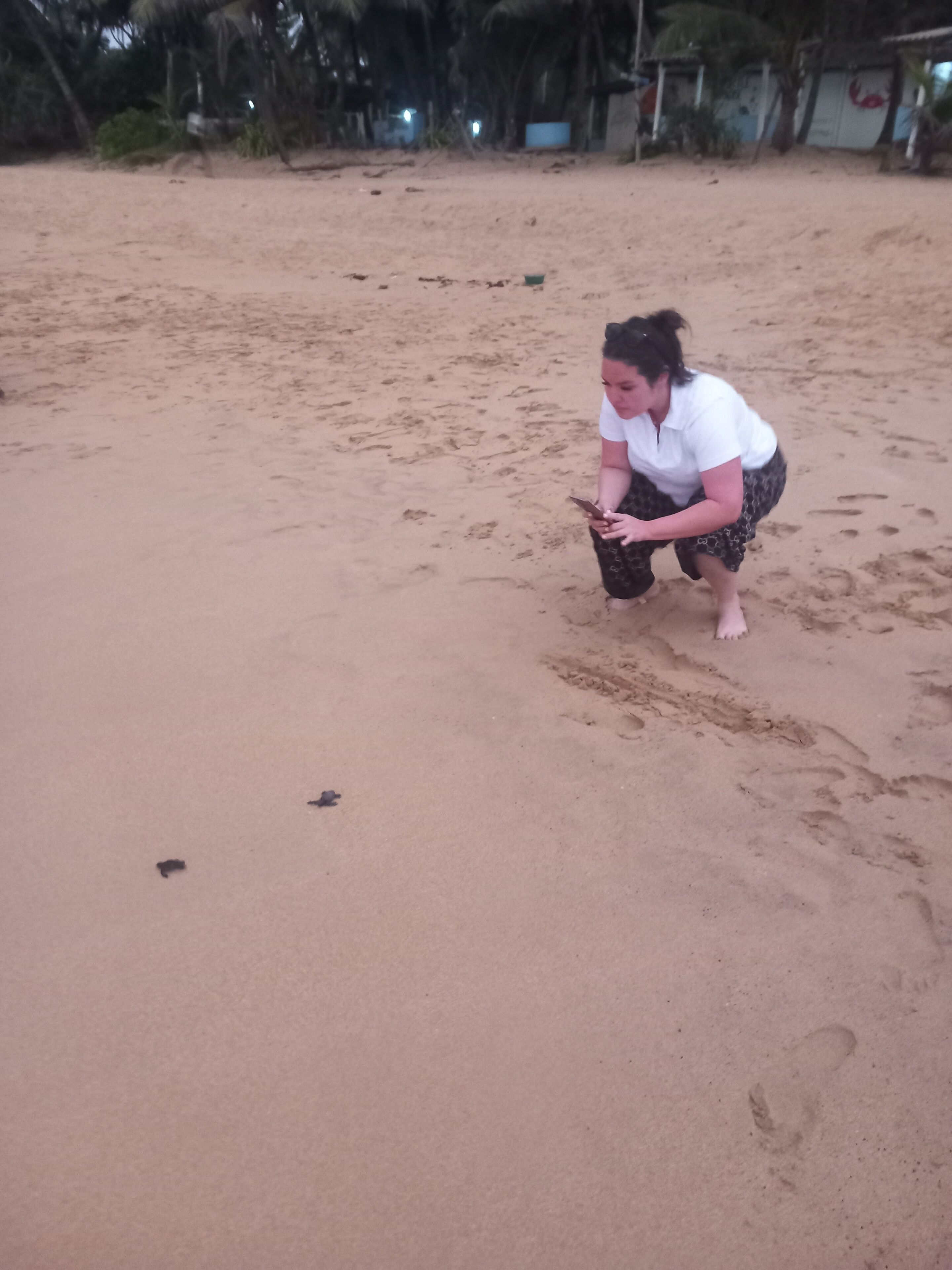 A person crouching on a sandy beach with footprints and a small object on the sand.