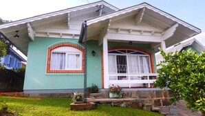 A house with a green facade, white trim, and a stone foundation. It features a front porch with a potted plant and a small garden area. - Residencial Encantos de Gramado (Gramado)