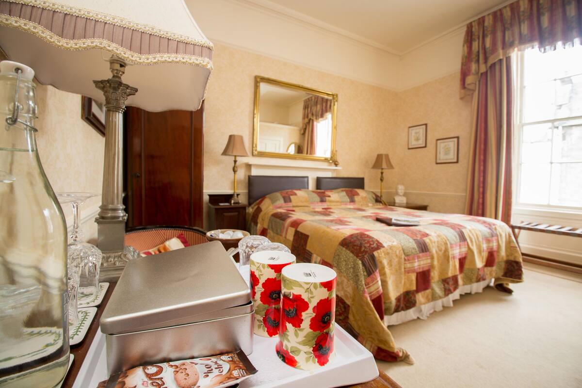 Habitación de hotel con cama, mesita de noche con lámpara, espejo y ventana con cortinas.