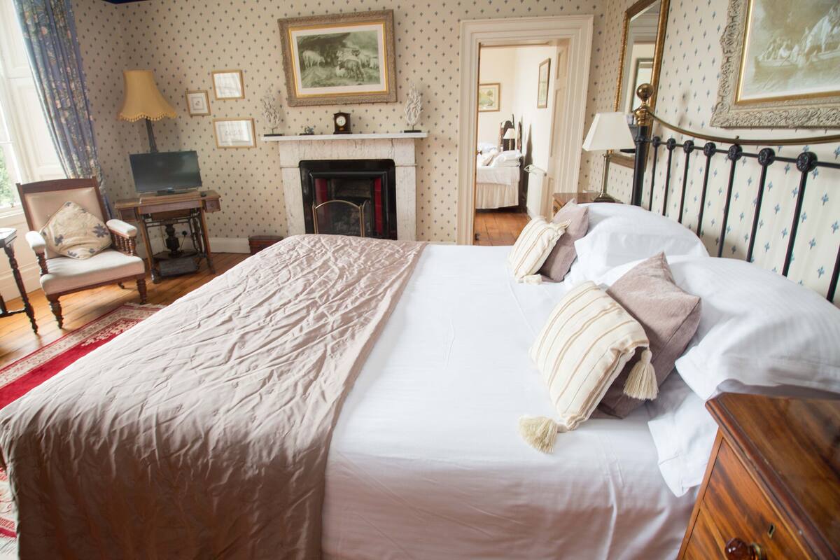 Un dormitorio ordenado con una cama grande, una silla de madera, un escritorio con lámpara, una chimenea y cuadros en la pared.
