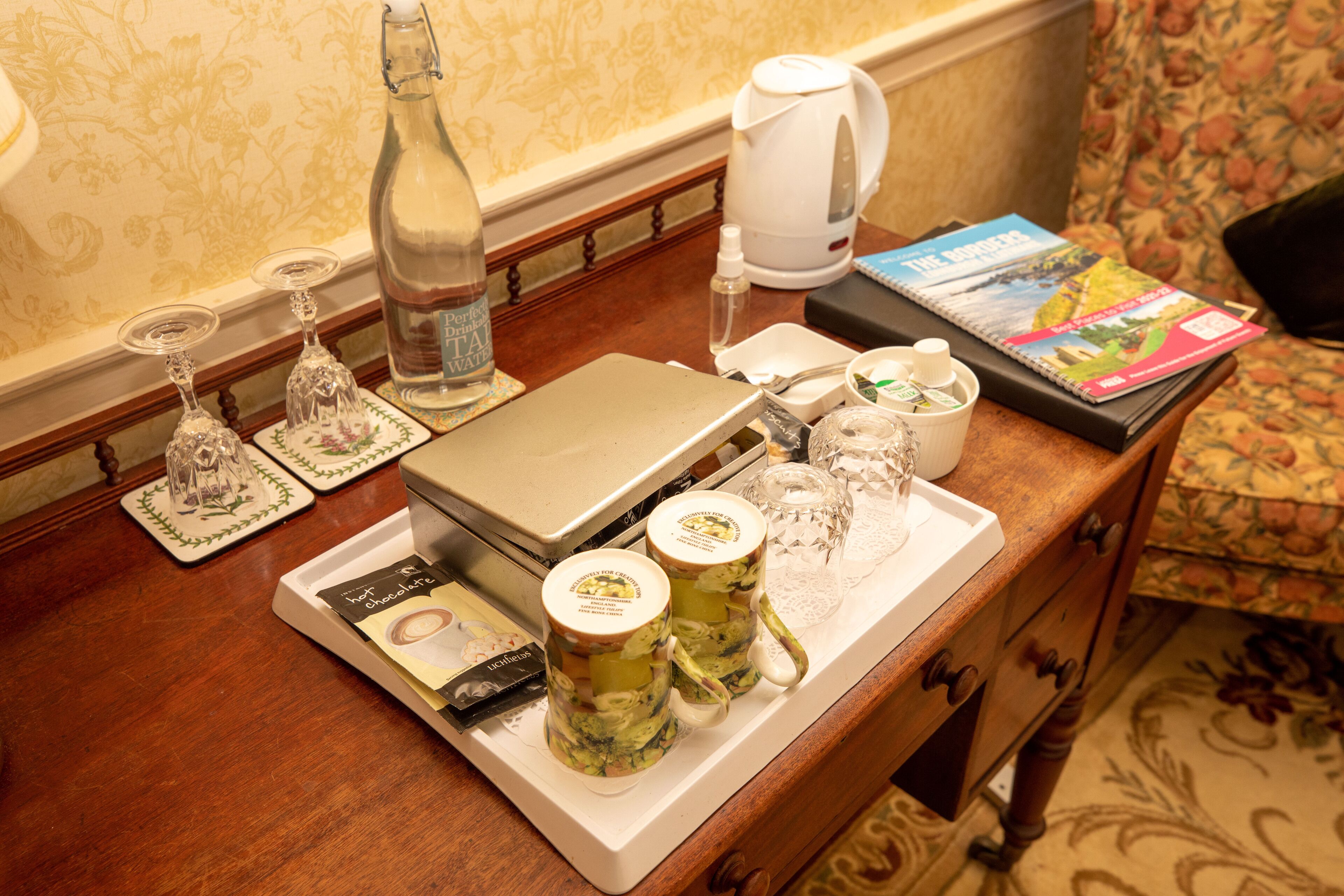 Een dienblad met een fles, glazen, een waterkoker en een boek op een houten tafel.