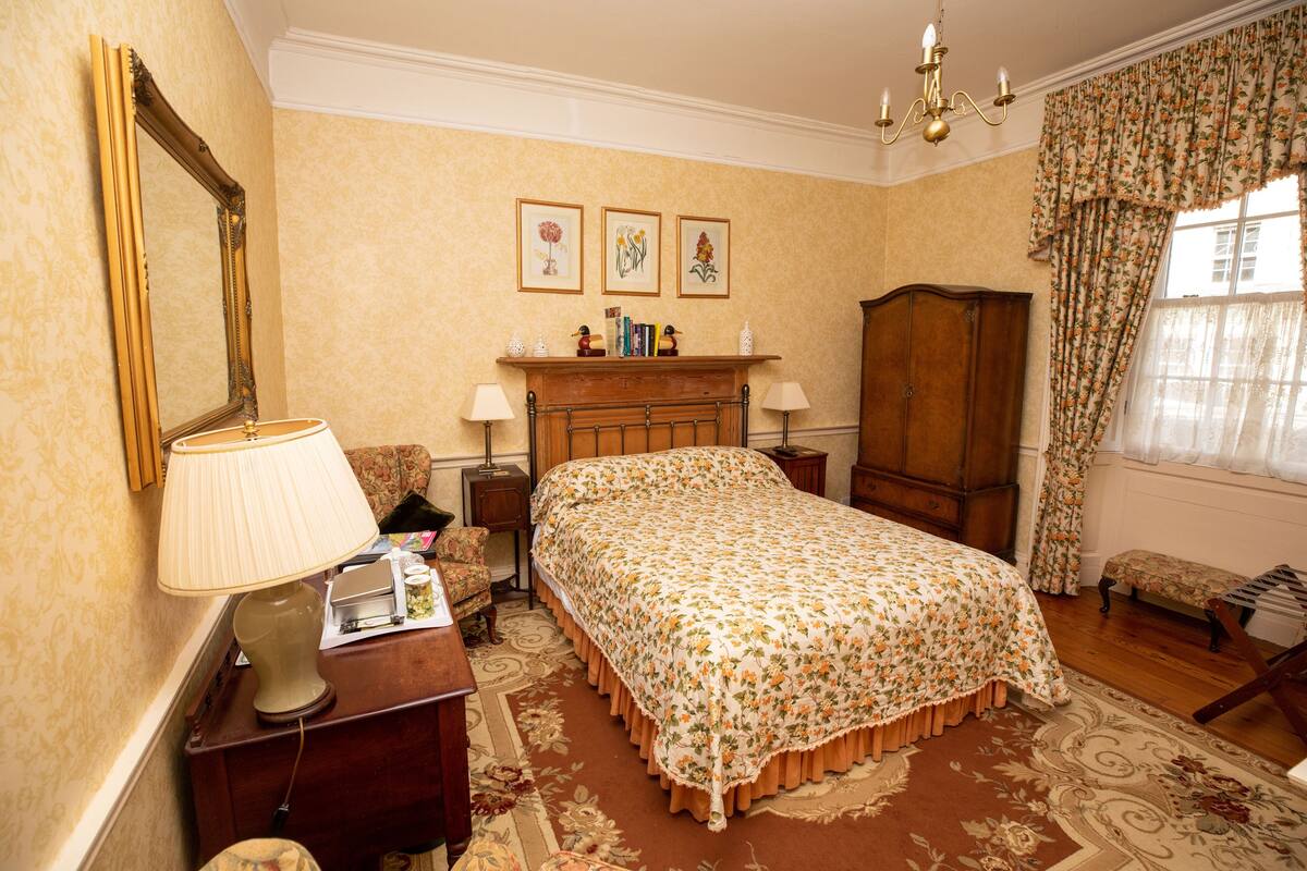 Un dormitorio con cama, mesita de noche, lámpara, silla, armario y espejo. Encima de la cama, hay tres cuadros enmarcados en la pared.
