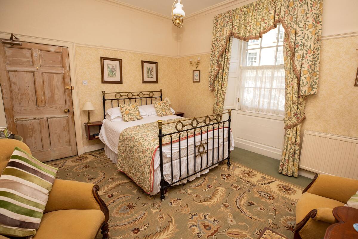 Un dormitorio con cama, una puerta de madera, una ventana con cortinas y una alfombra estampada.