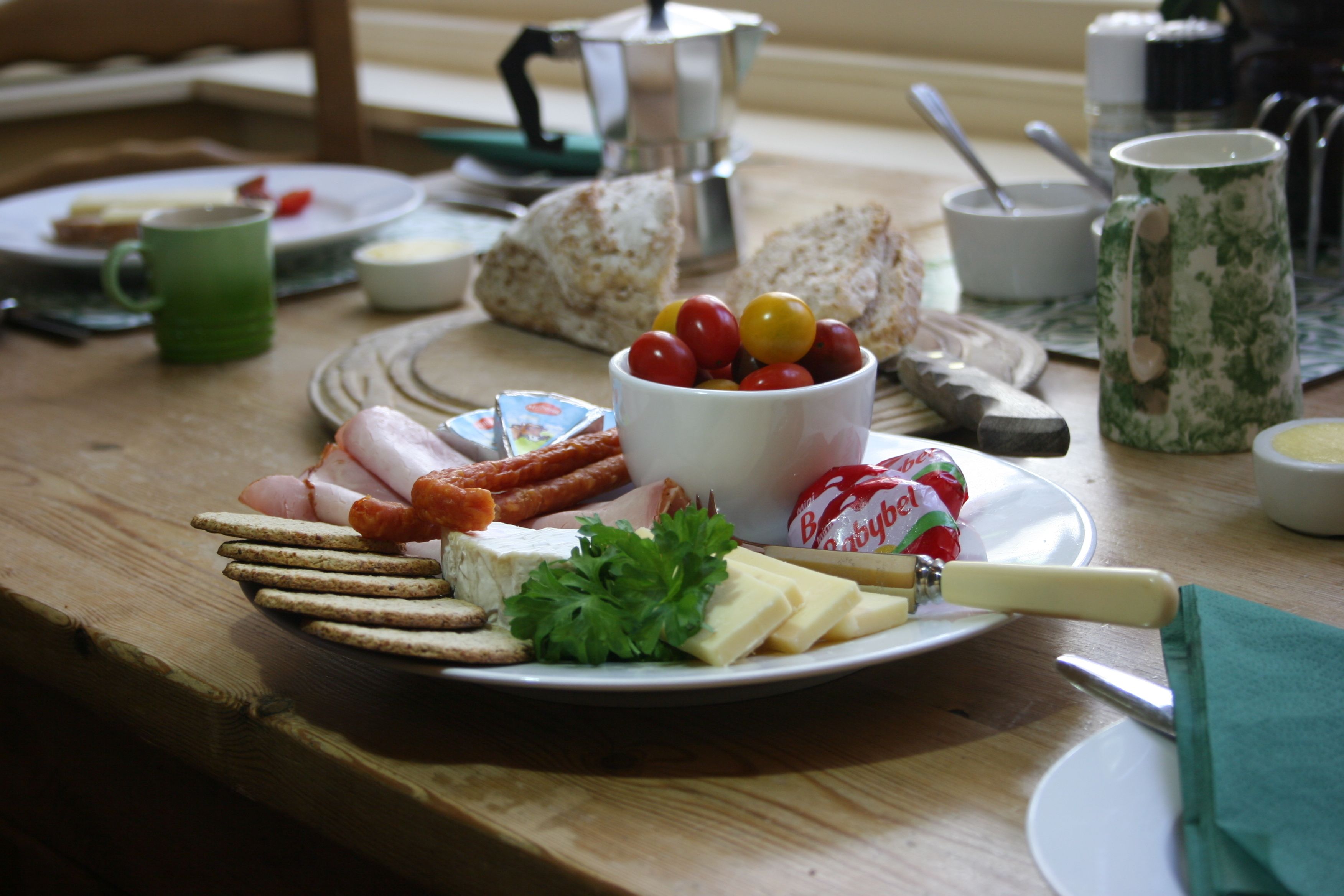 A breakfast setting with a plate of cheese, crackers, and tomatoes, a green mug, and a coffee pot on a wooden table.