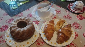 Taula amb un plat de postre que conté un pastís bundt i dos croissants, una teiera, i una tassa amb una cullera sobre un mantell amb un dibuix floral.