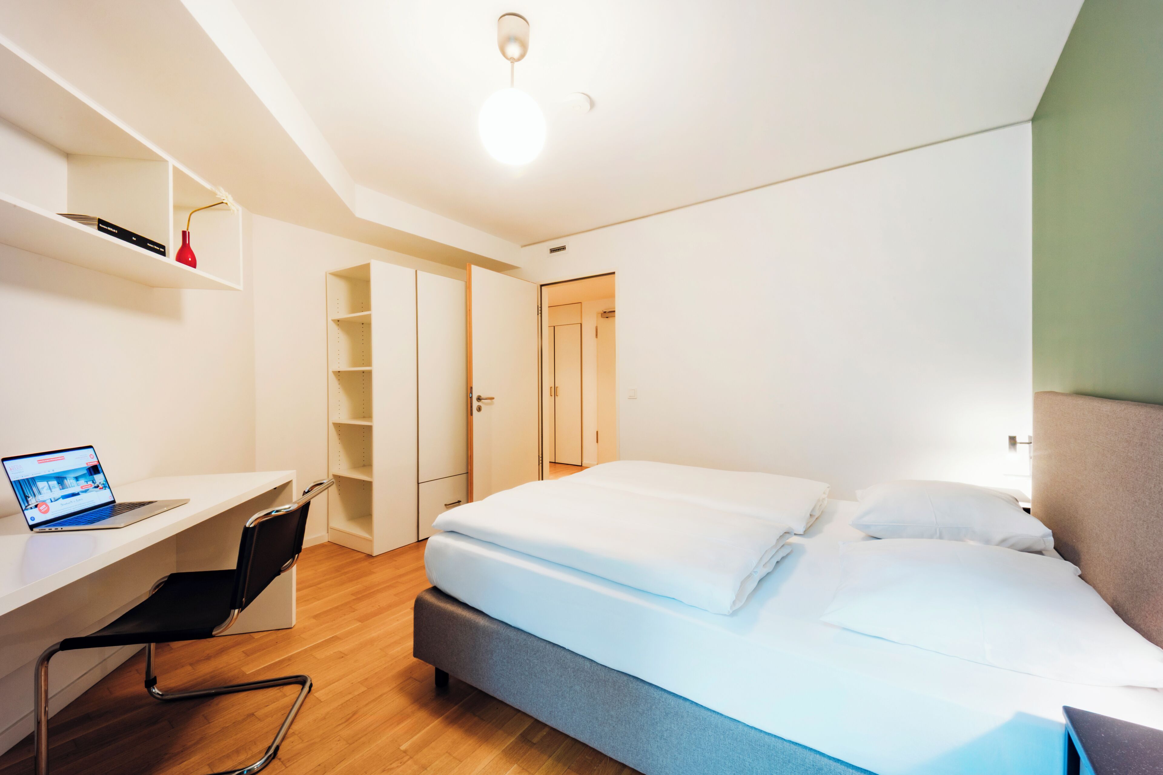 A modern hotel room with a bed, a desk with a laptop, a chair, and built-in wardrobes.