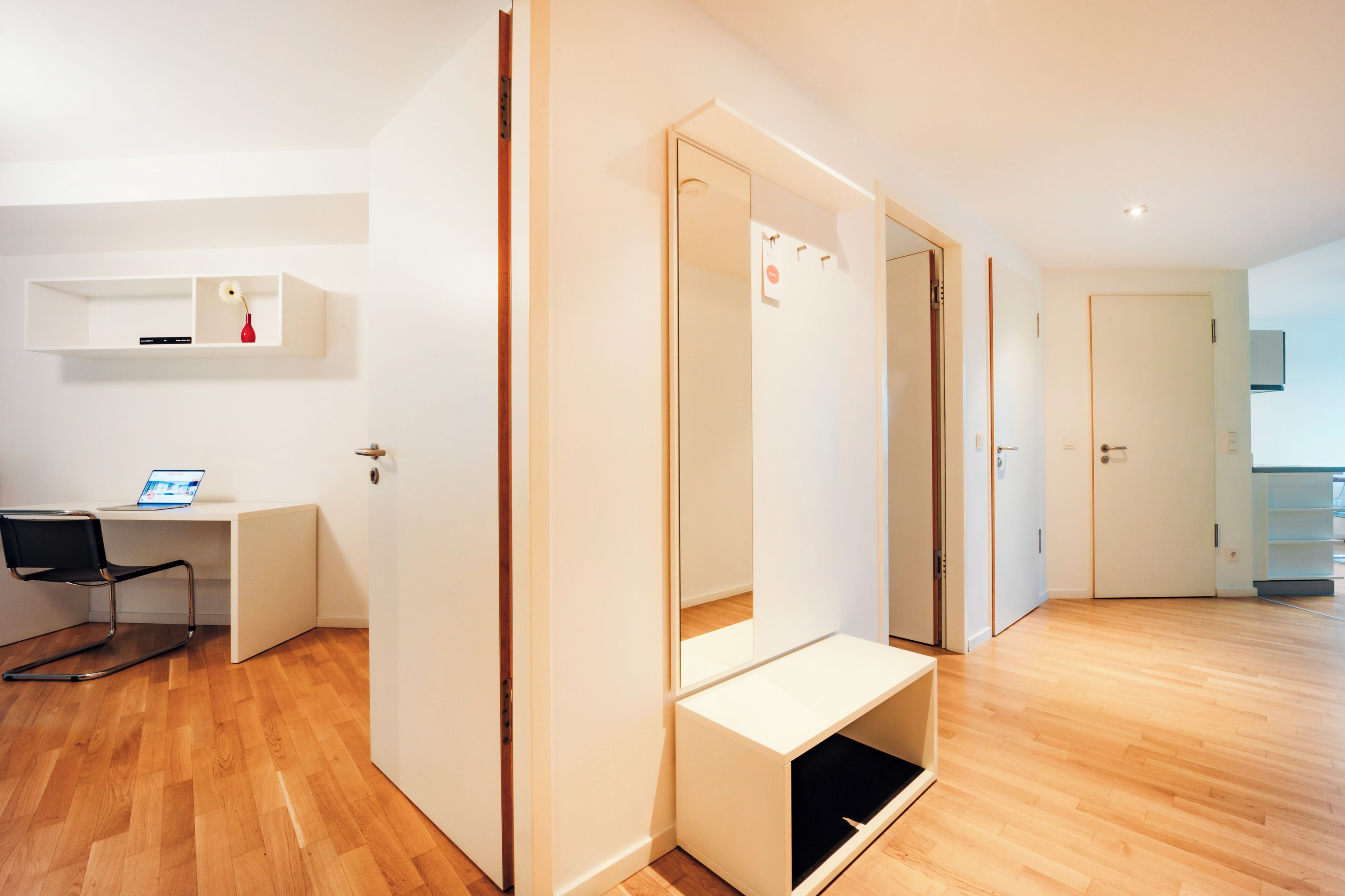Ein modernes Hotelzimmer mit Holzboden, Schreibtisch, Laptop, Stuhl und Kleiderschrank mit Spiegel.
