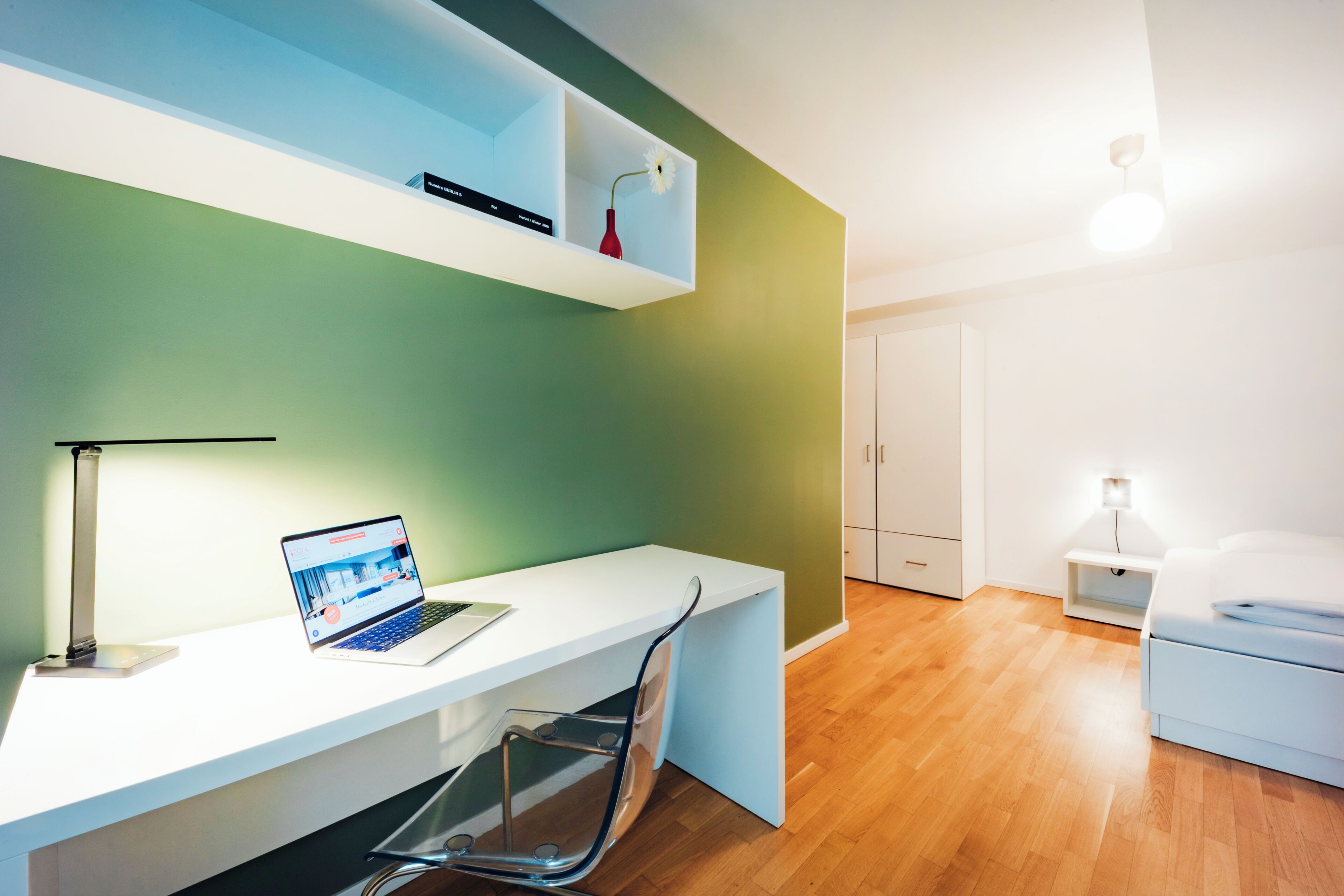 A modern room with a white desk, a laptop, and a chair. There is a shelf with a red vase and a white flower. The room has green walls and a wooden floor.
