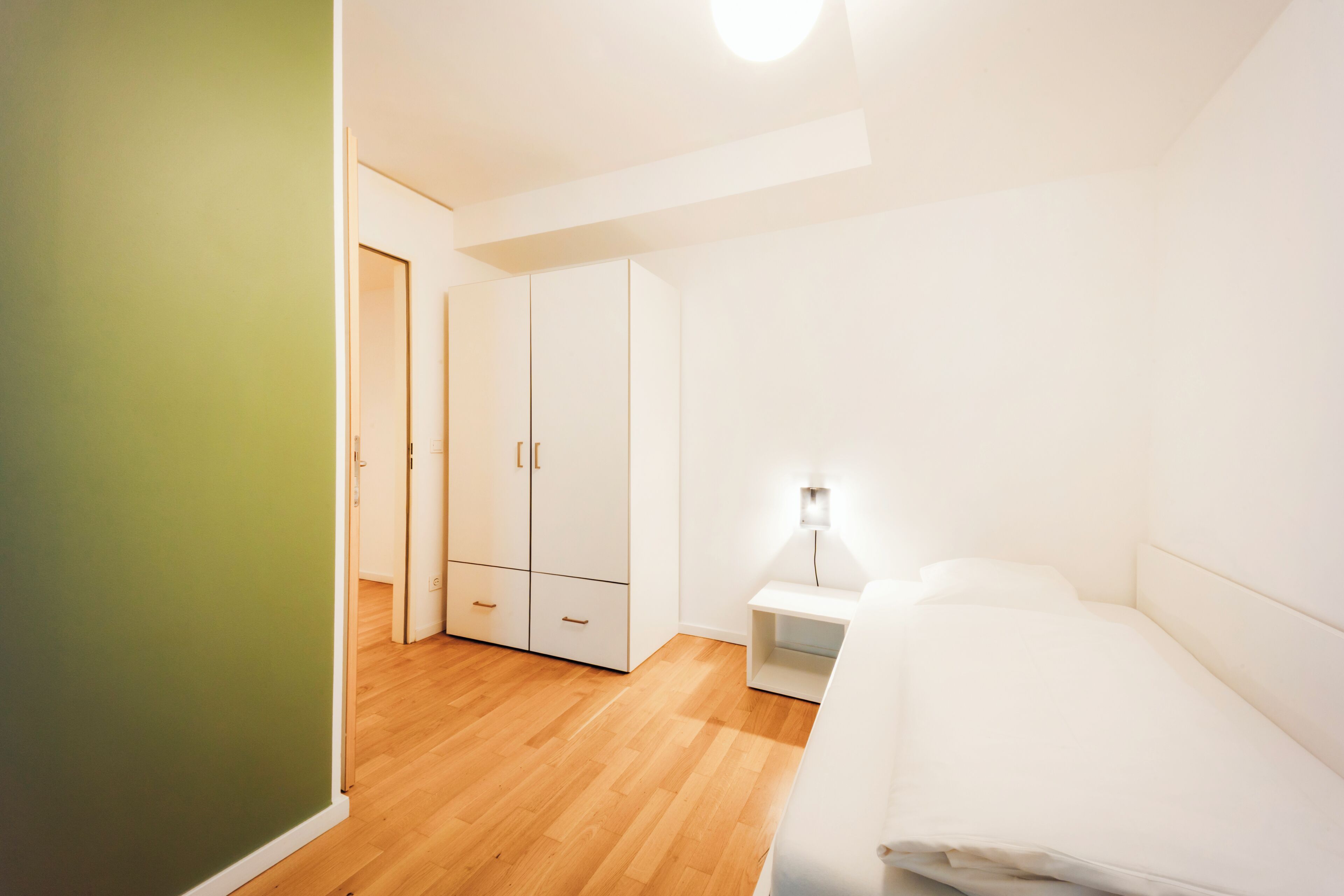 Quarto minimalista com uma cama, mesa de cabeceira, guarda-roupa e uma parede verde.