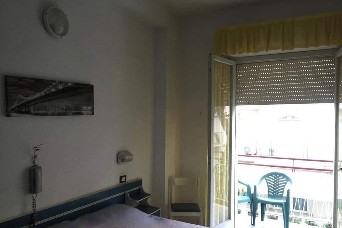 A hotel room with a bed, a telephone, a small table, a chair, and a balcony with chairs.