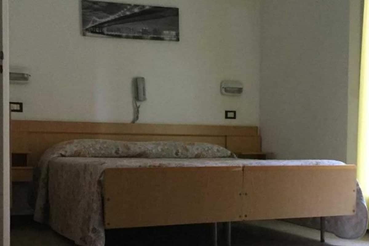A single bed with a headboard, a wall-mounted light, a framed picture, and a window with a curtain.