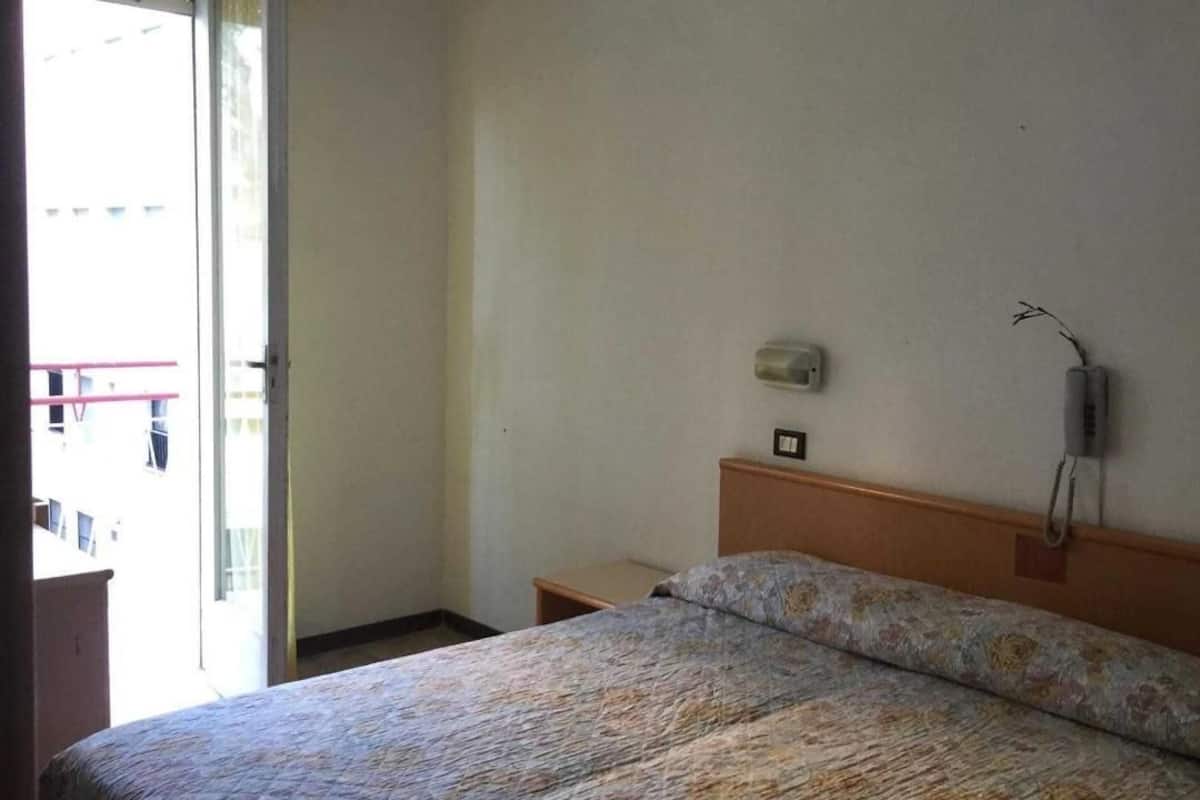 A hotel room with a bed, a bedside table, a wall-mounted phone, and a window with a view of the outside.