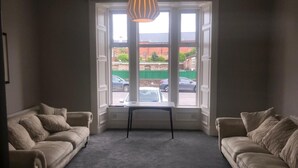 A room with two beige sofas, a small table, and a large window with a view of the street and parked cars. - Aberdour Guest House (Dumfries)