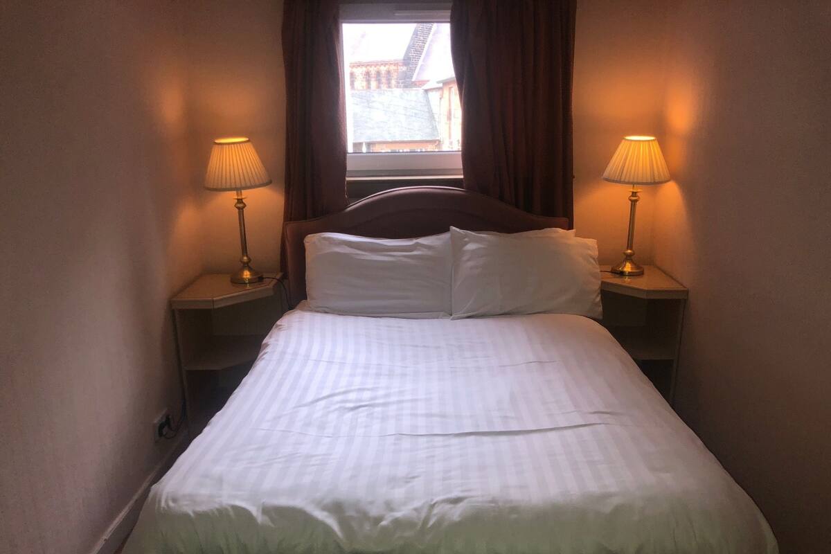 A hotel room with a single bed, two bedside lamps, and a window with curtains.