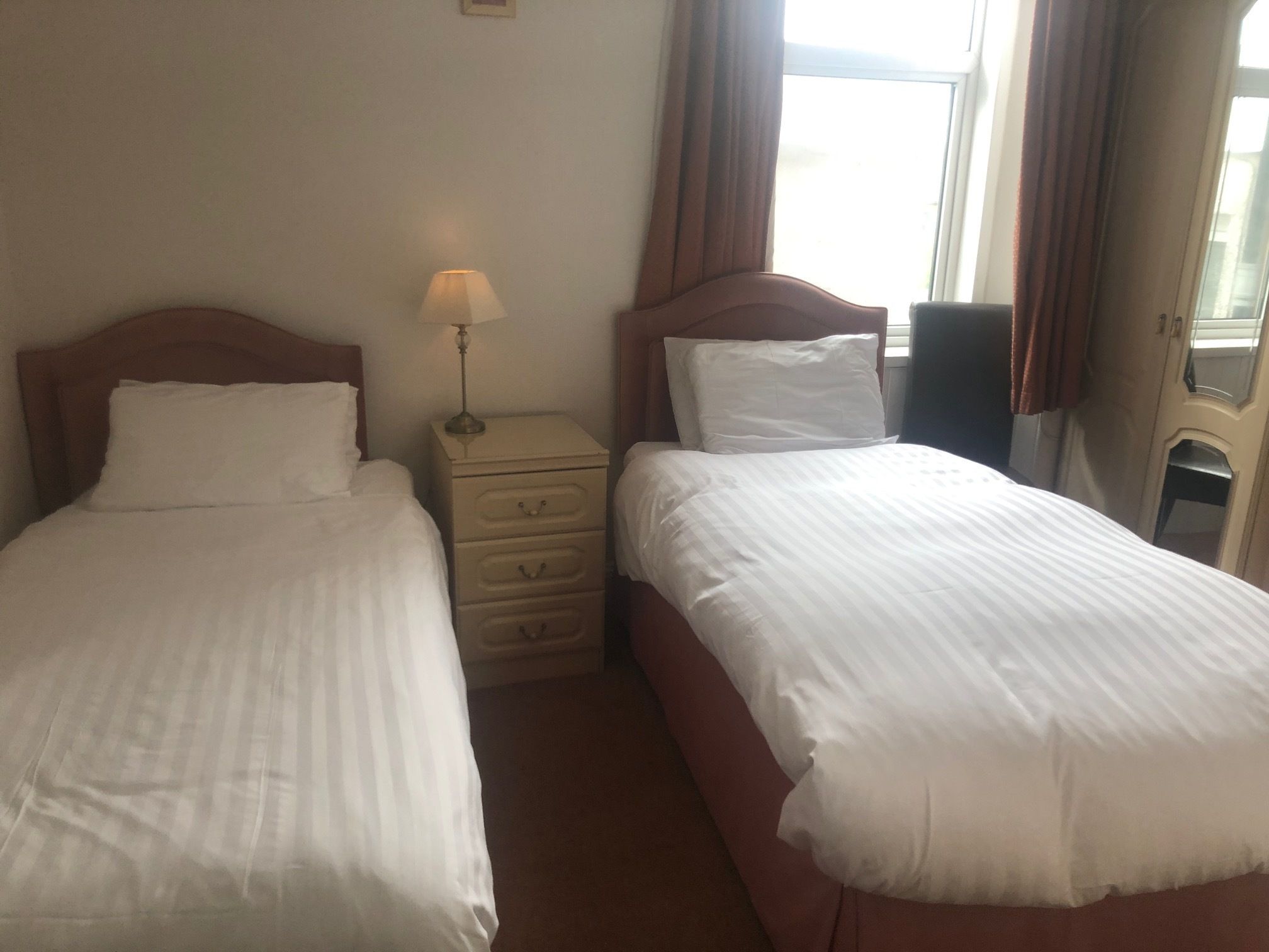 Habitación de hotel con dos camas individuales, una mesita de noche con lámpara y una ventana con cortinas.