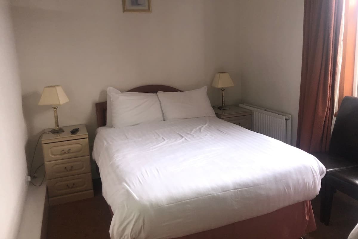 A neatly made bed with white linens, two bedside tables with lamps, a radiator, and a chair.