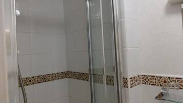 Área de duche com divisória de vidro, paredes revestidas a azulejo branco e um rodapé decorativo em azulejo.