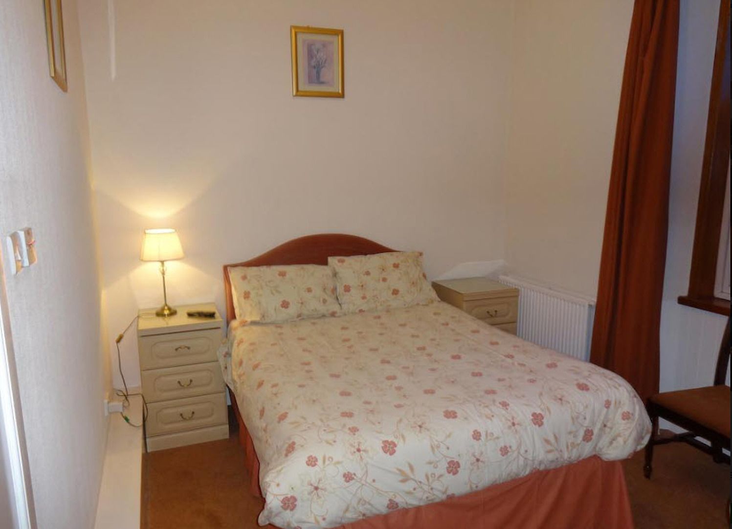 Een slaapkamer met een bed, nachtkastje, lamp, radiator en een stoel.