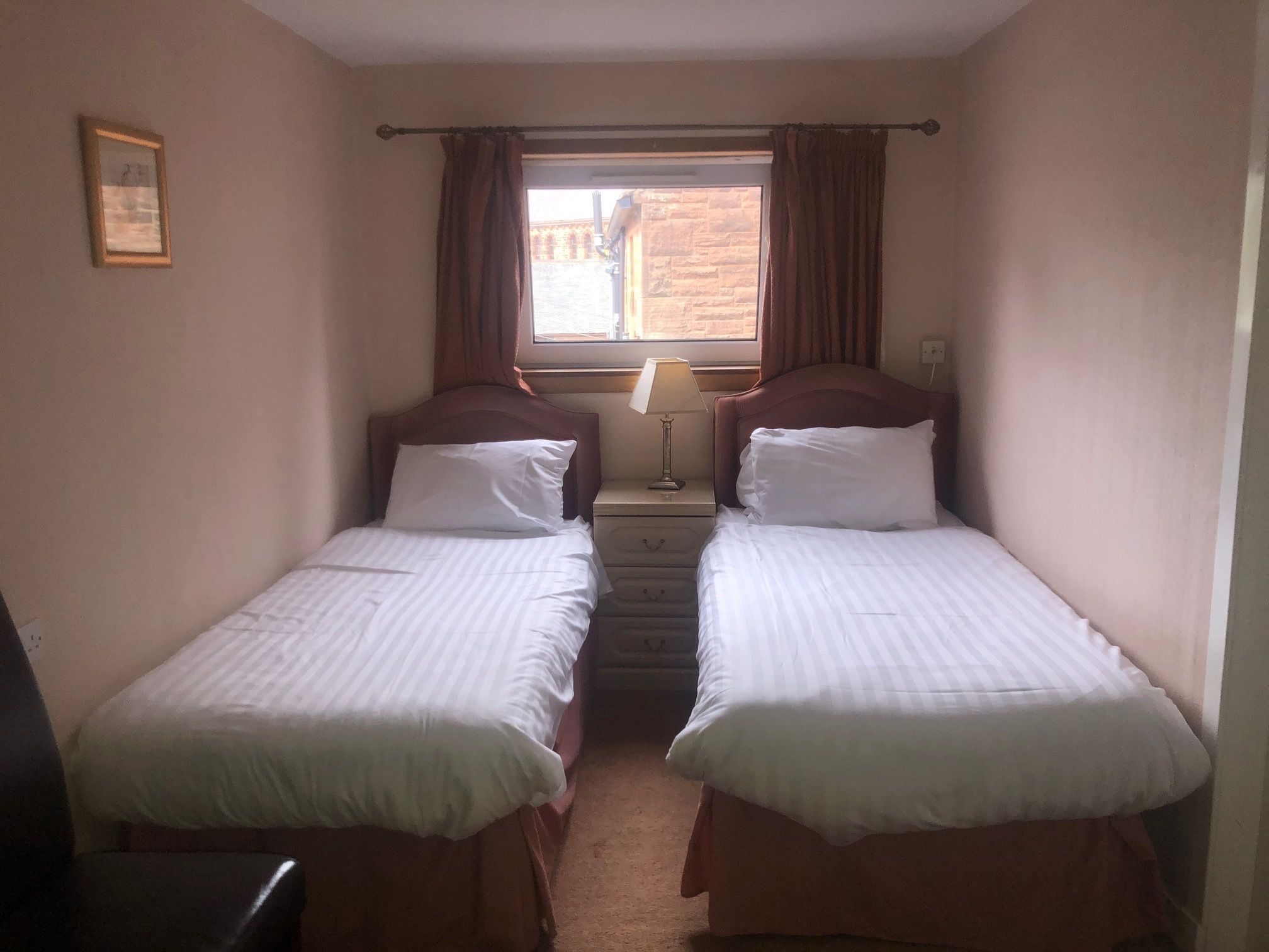 Een tweepersoonskamer met twee eenpersoonsbedden, een nachtkastje met lamp, een raam met gordijnen en een lijst met een afbeelding aan de muur.