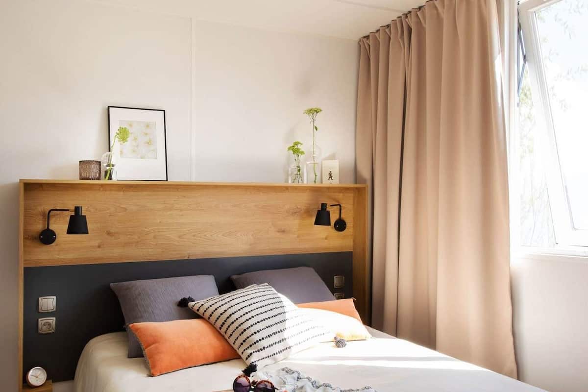 Una cama bien hecha con cabecera de madera, varios almohadones y una mesita de noche con lámpara.