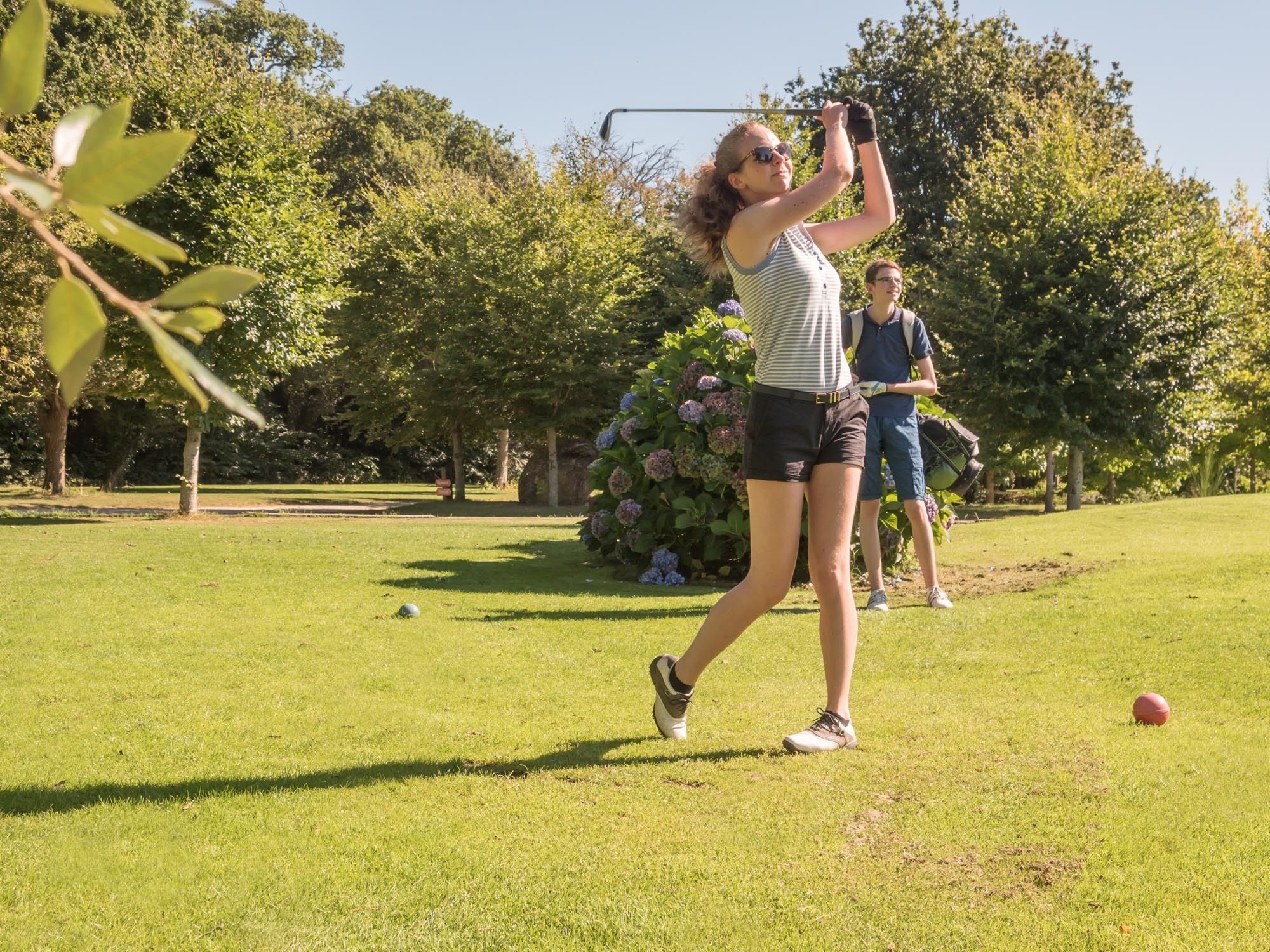 Eine Person spielt Golf auf einem gepflegten Rasenplatz, im Hintergrund Bäume und Blumen.
