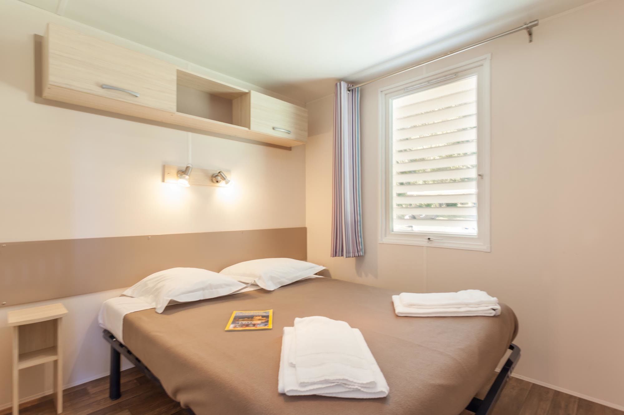 Hotellihuone, jossa on sänky, yöpöytä ja ikkuna, jossa on sälekaihtimet.