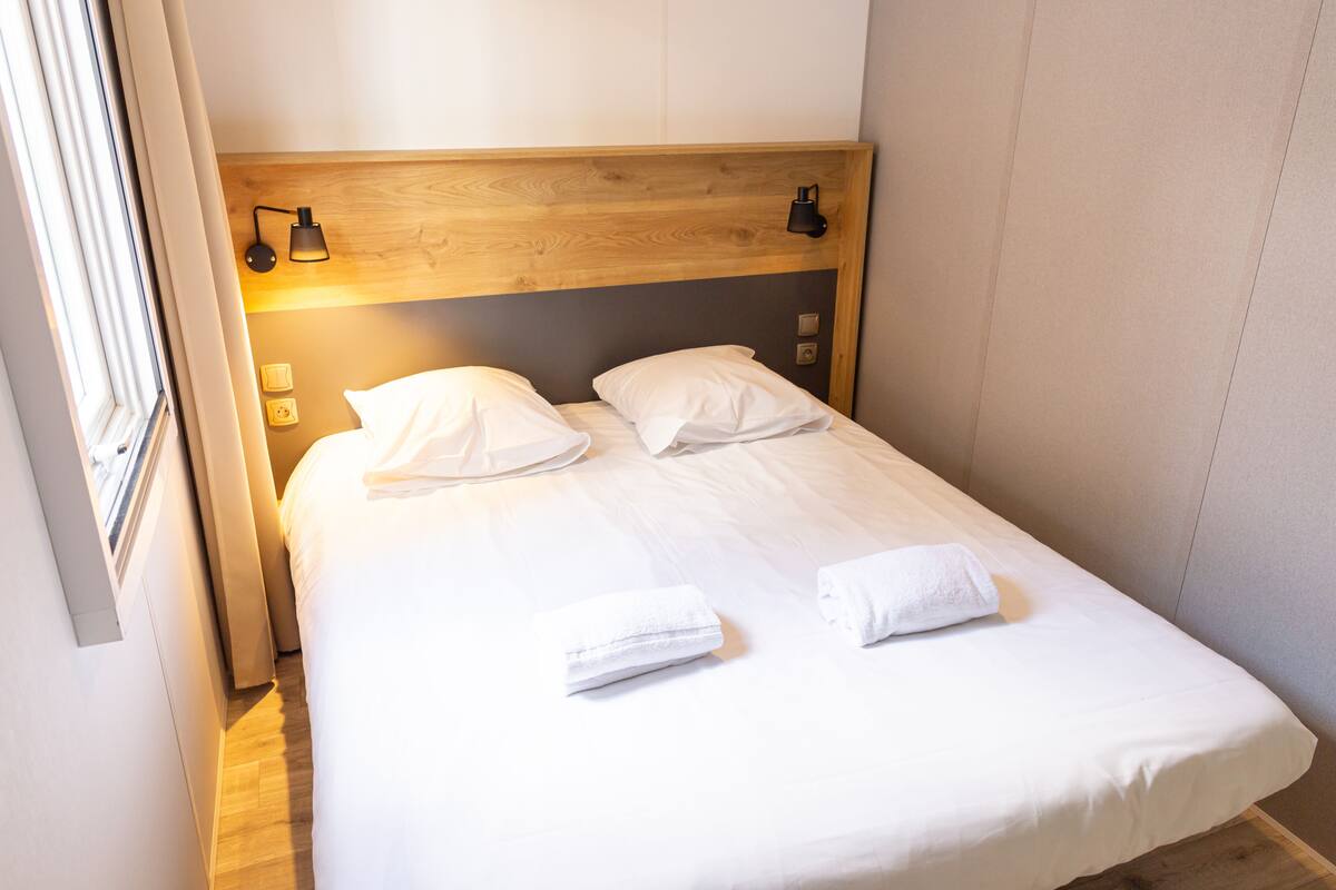 Una cama bien hecha con sábanas blancas y dos toallas, cabecera de madera y lámparas de noche.