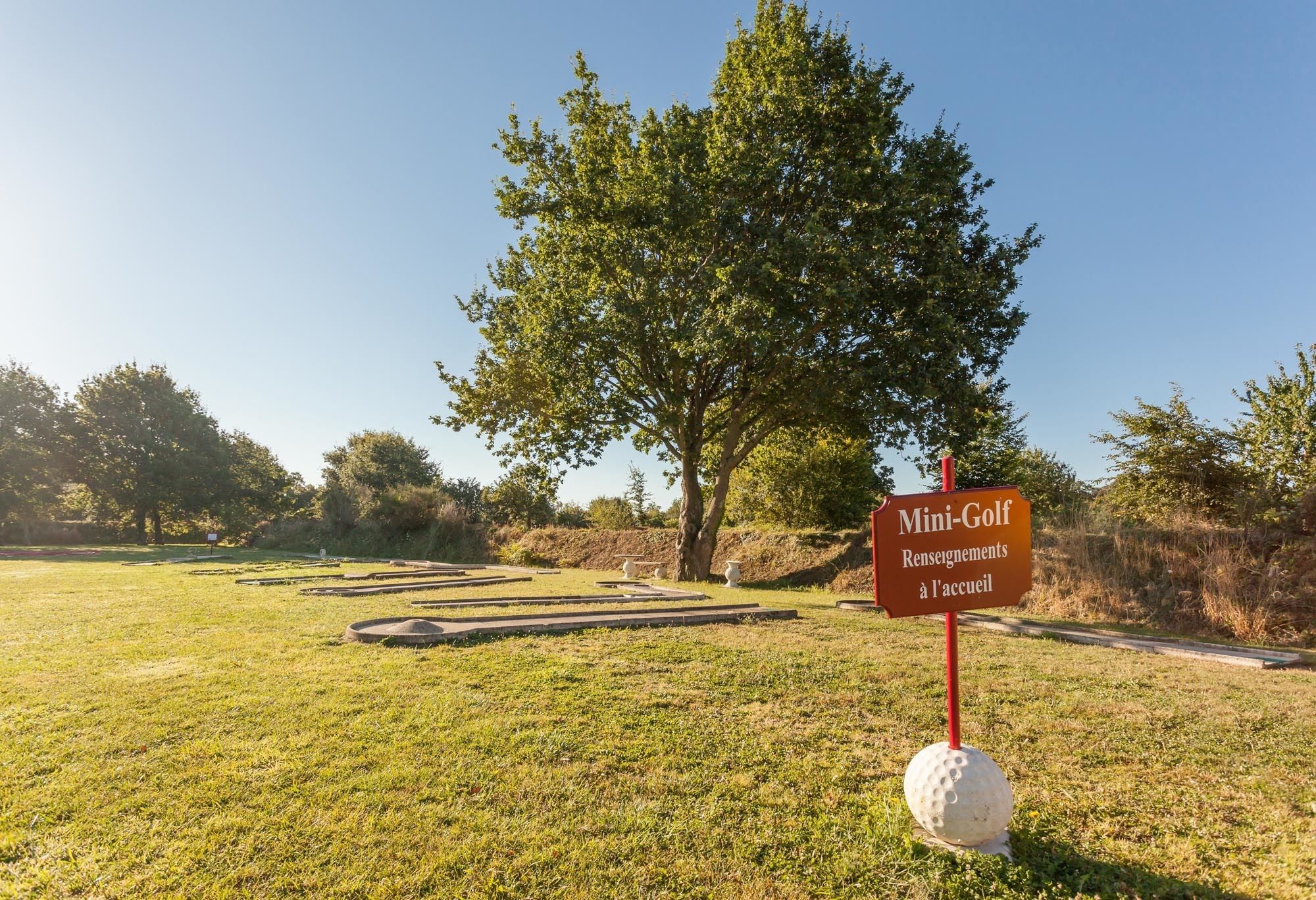 Naamplaatje voor een minigolfbaan, met een golfbal aan de voet, gelegen in een grasrijk gebied met een grote boom op de achtergrond.