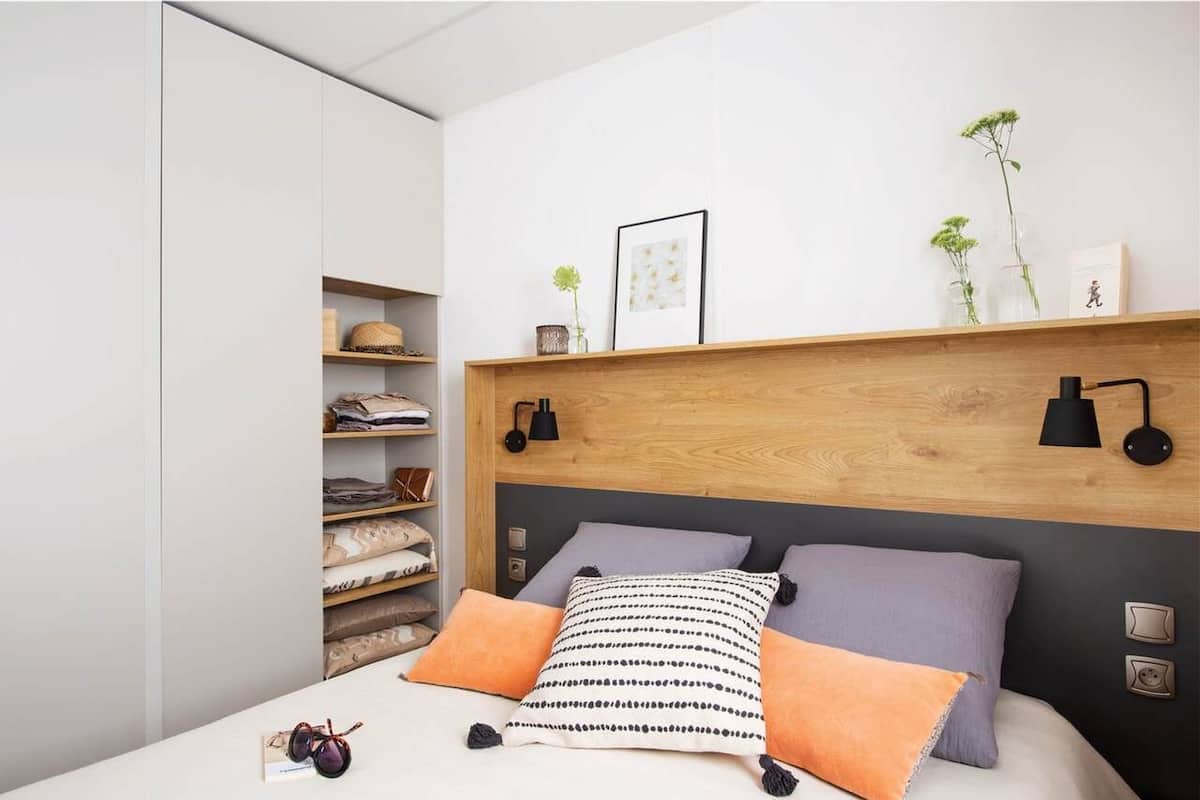 Un dormitorio moderno con cabecera de madera, cama con almohadas estampadas, mesita de noche con lámpara y estante con objetos decorativos.