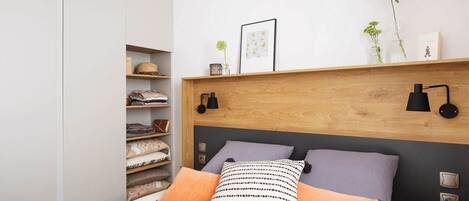 Une chambre moderne avec une tête de lit en bois, un lit agrémenté d’oreillers à motifs, une table de chevet avec une lampe et une étagère ornée d’objets décoratifs.