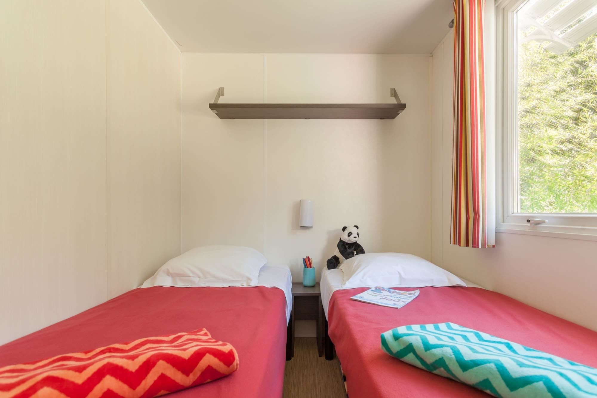 Ein kleines Zimmer mit zwei Betten, einem Regal darüber, einem Panda-Plüschtier und einem Fenster mit Vorhängen.