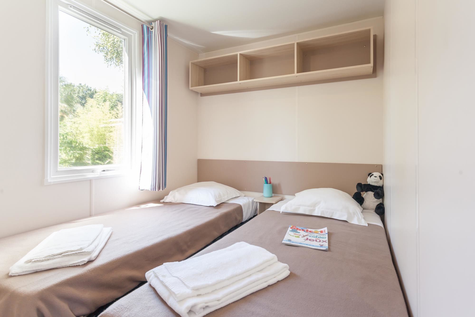 Ein Hotelzimmer mit zwei Einzelbetten, einem Fenster mit Vorhängen, einem Regal über den Betten und einem Plüschpanda auf einem Bett.