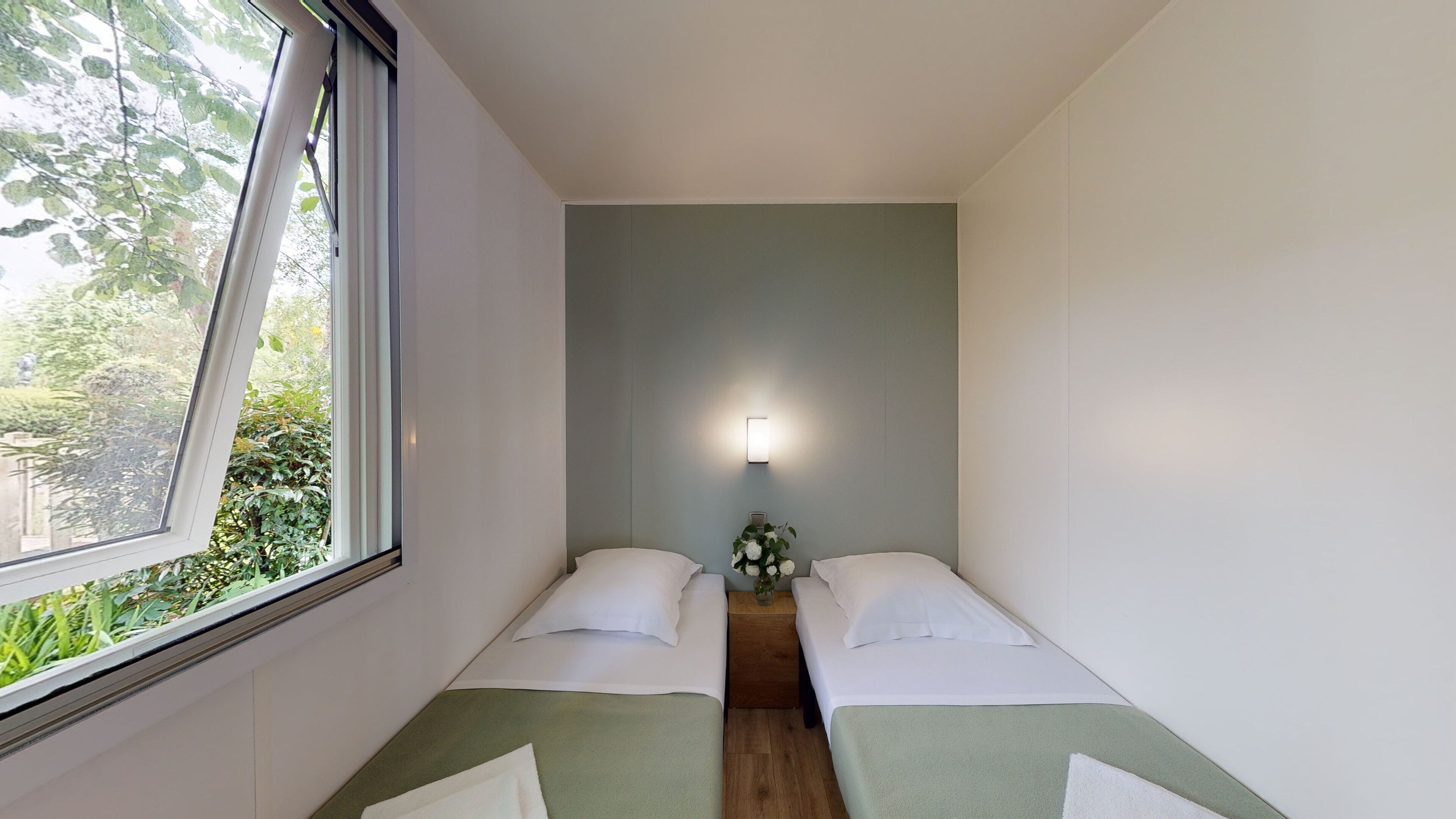 Een kleine, schone kamer met twee bedden, een nachtkastje met een bloevas en een raam met uitzicht op groen.