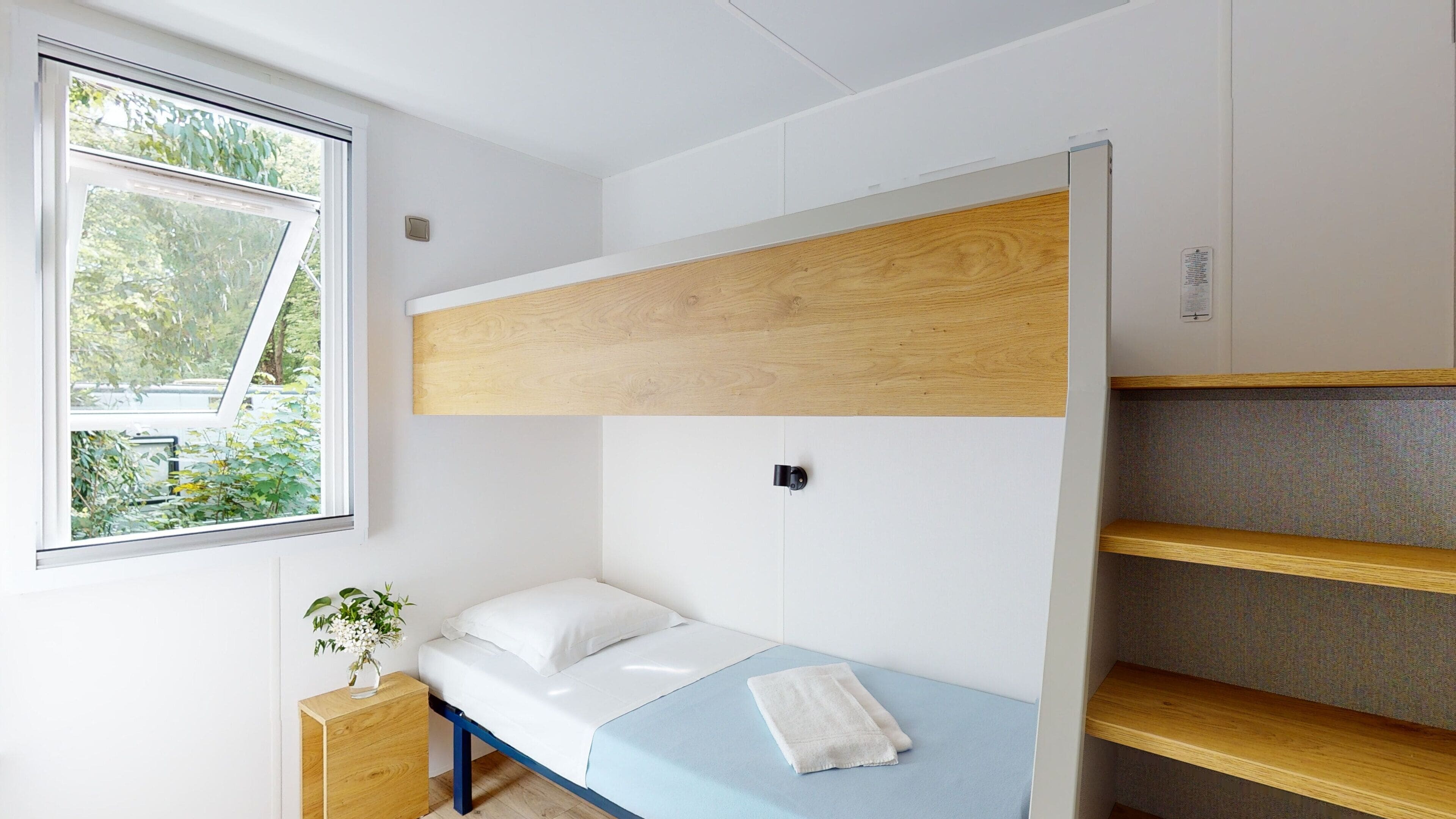 Une petite chambre avec un lit superposé, une petite table de chevet agrémentée d’une plante et une fenêtre donnant sur un paysage verdoyant.