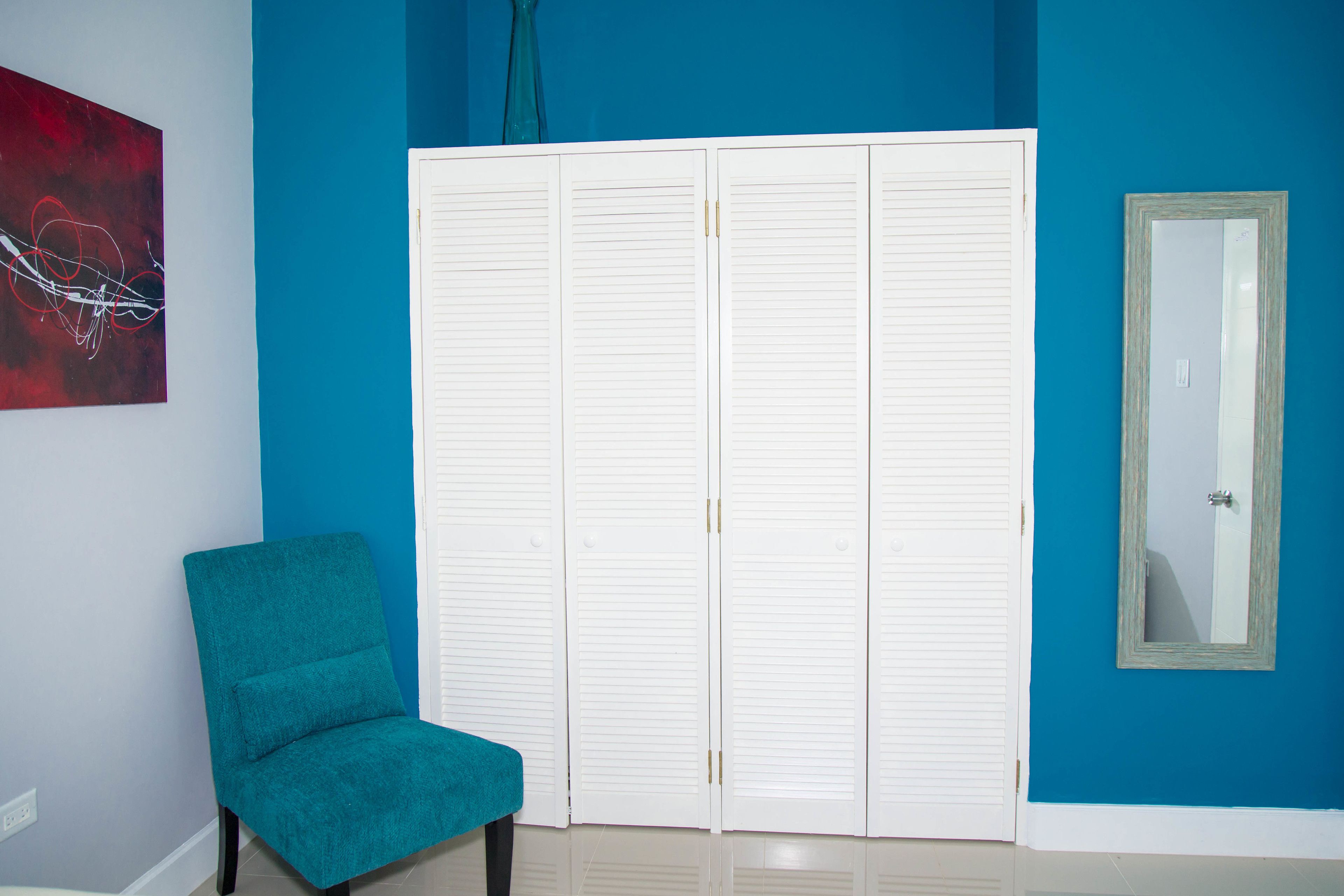Una habitación con paredes verde azulado, un armario blanco con puertas correderas, una silla verde azulado y un espejo en la pared.