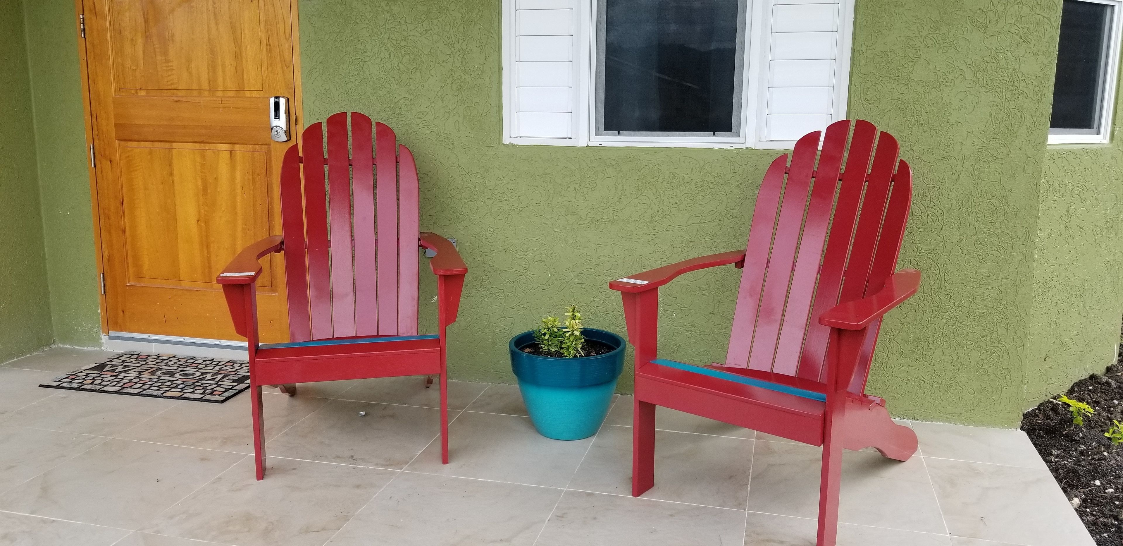 Two red Adirondack chairs outside a house with a green textured wall, a wooden door, and a window with white shutters.