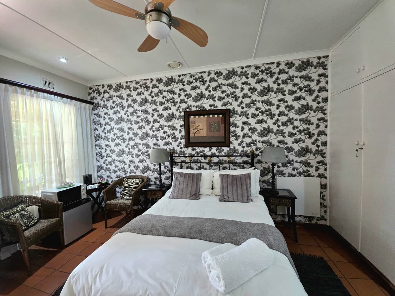 A bedroom with a bed, two nightstands, a ceiling fan, a framed picture, a chair, a desk, and a window with curtains.