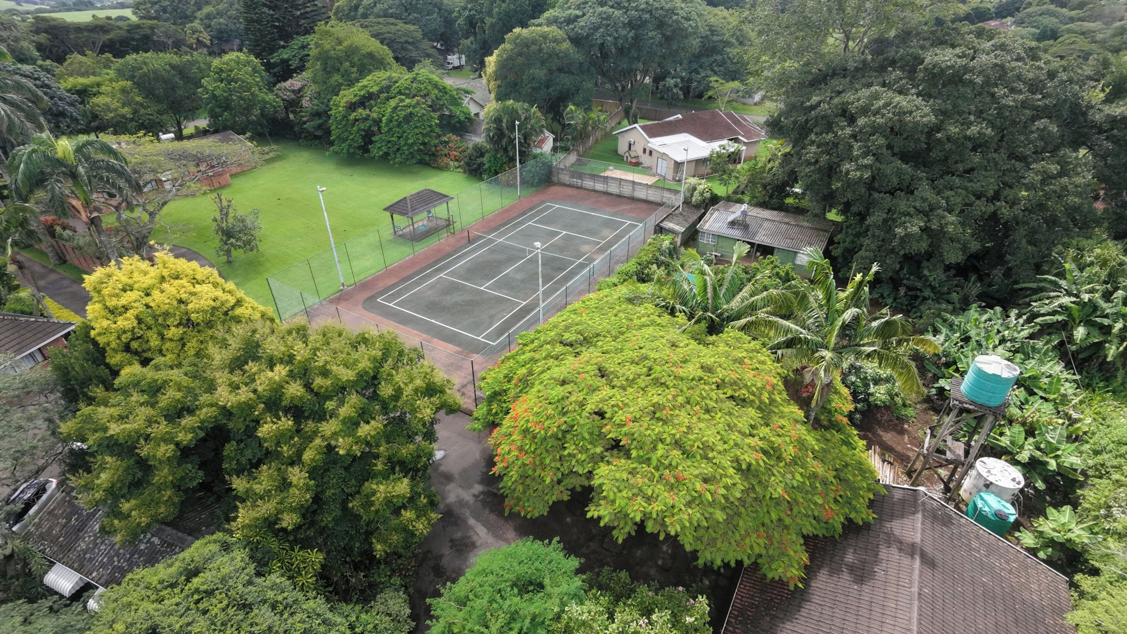 An aerial view of a residential area with a tennis court, lush greenery, and a small house.