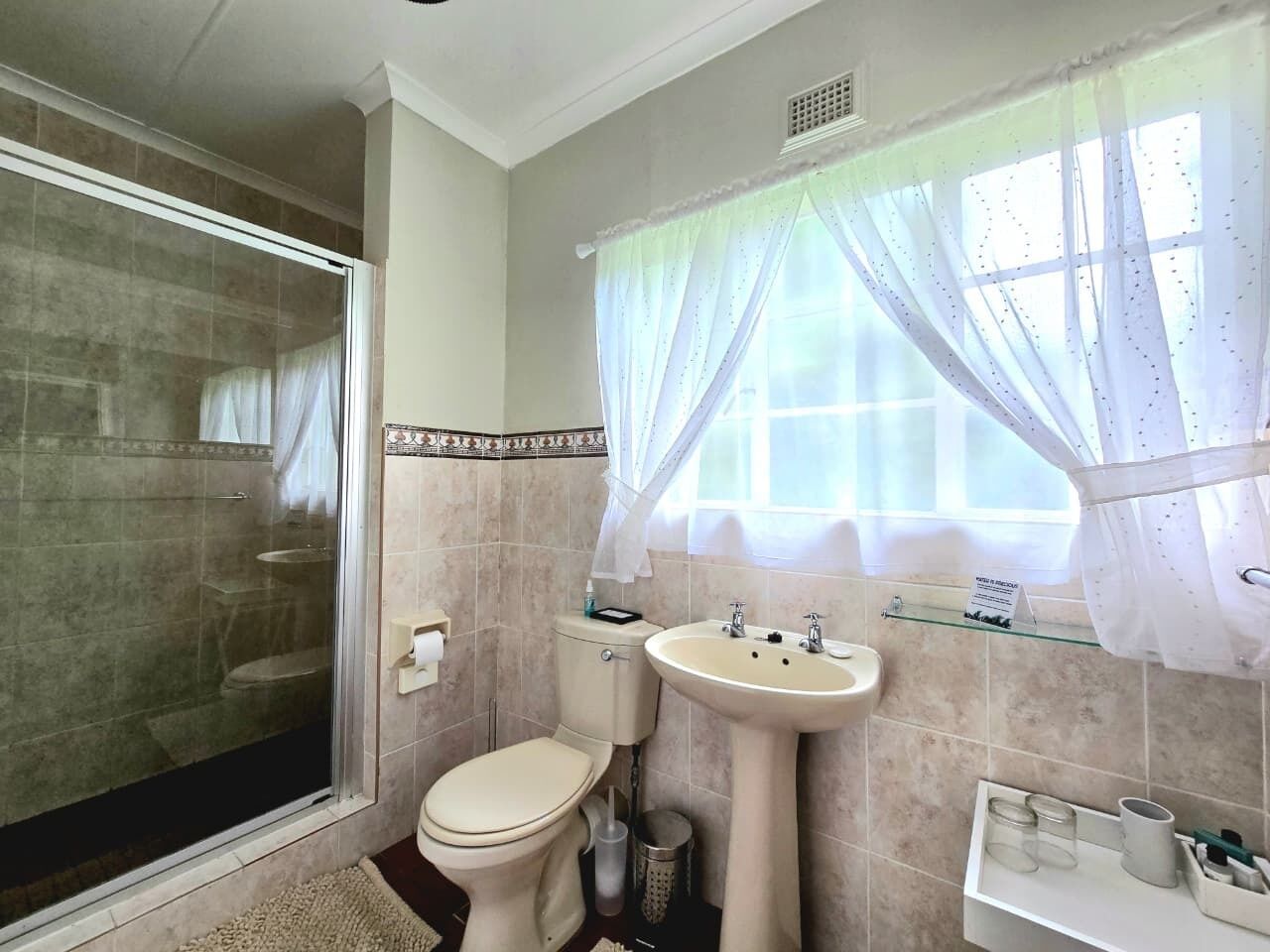 A bathroom with a shower, toilet, sink, and a window with sheer curtains.