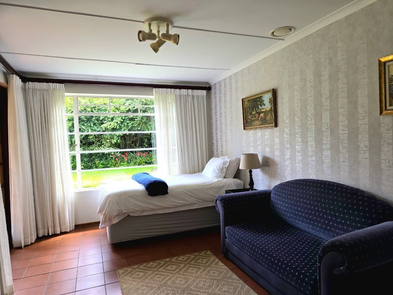 A bedroom with a bed, a blue sofa, a window with curtains, and a painting on the wall.