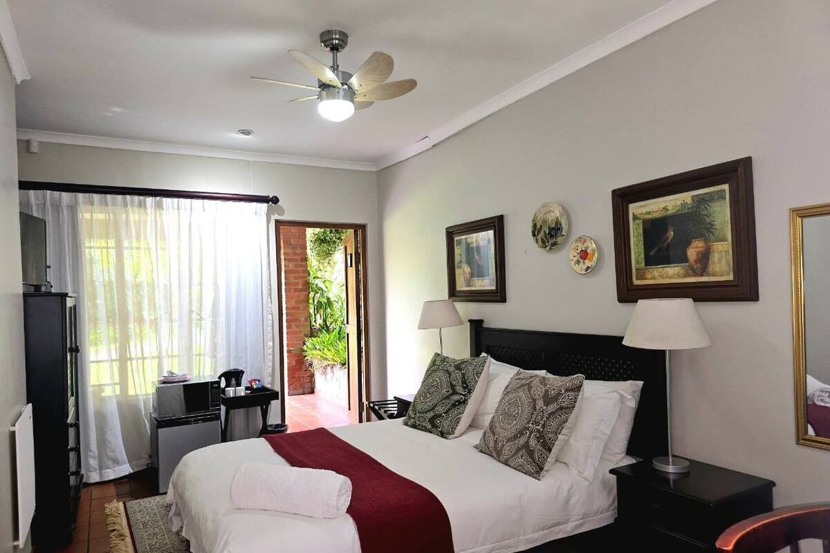 A bedroom with a bed, a desk, a chair, a TV, a ceiling fan, and a door leading to a balcony.