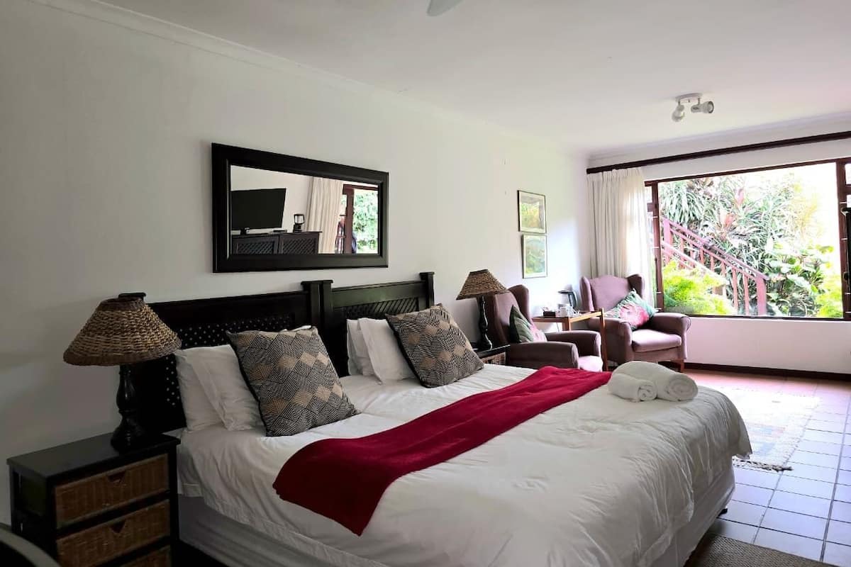 A bedroom with a bed, two bedside tables, a ceiling fan, a large window with a view of greenery, and a mirror on the wall.