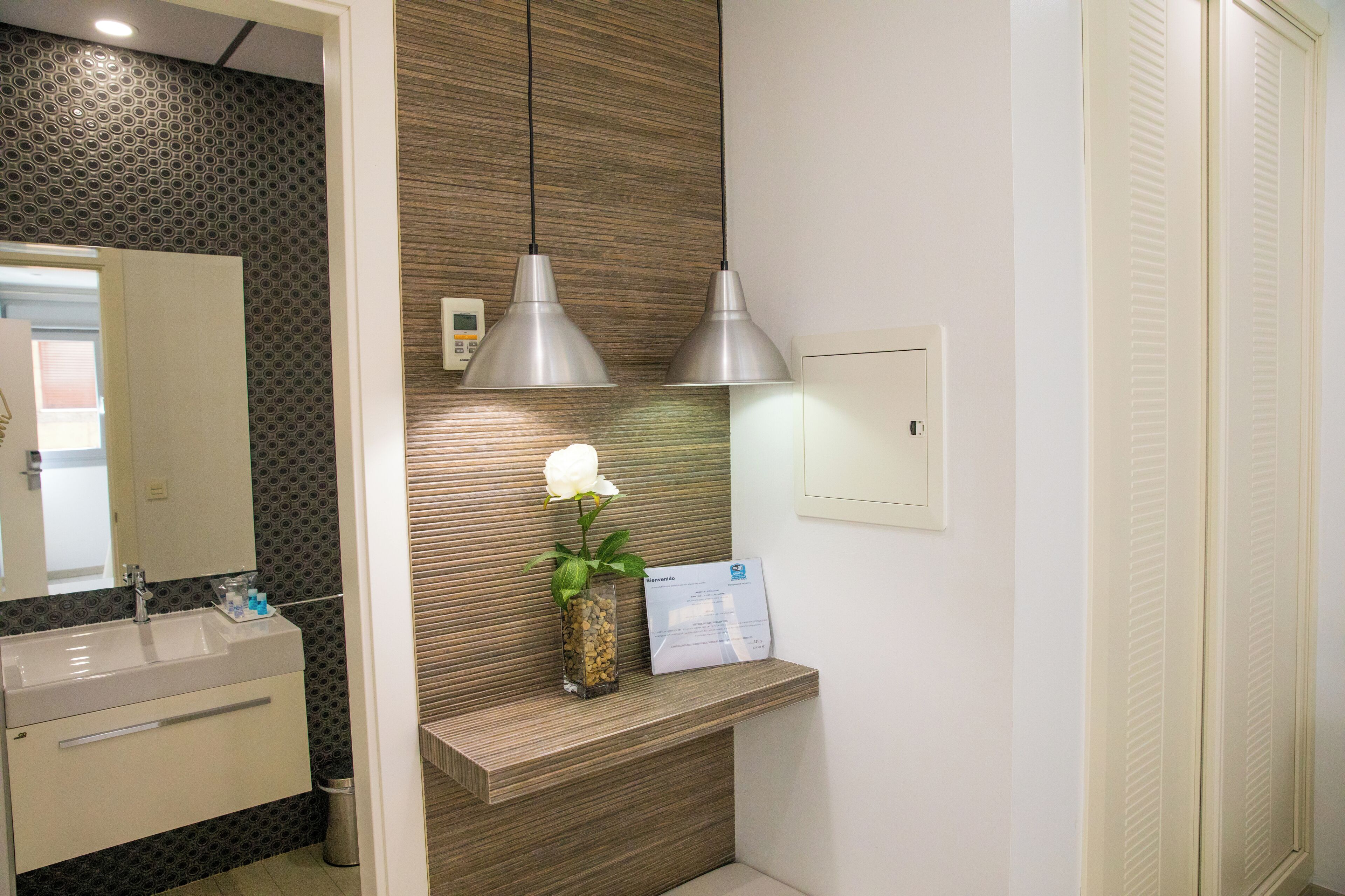A bathroom with a sink, mirror, vanity, two pendant lights, a vase with a flower, and a sign on the wall.